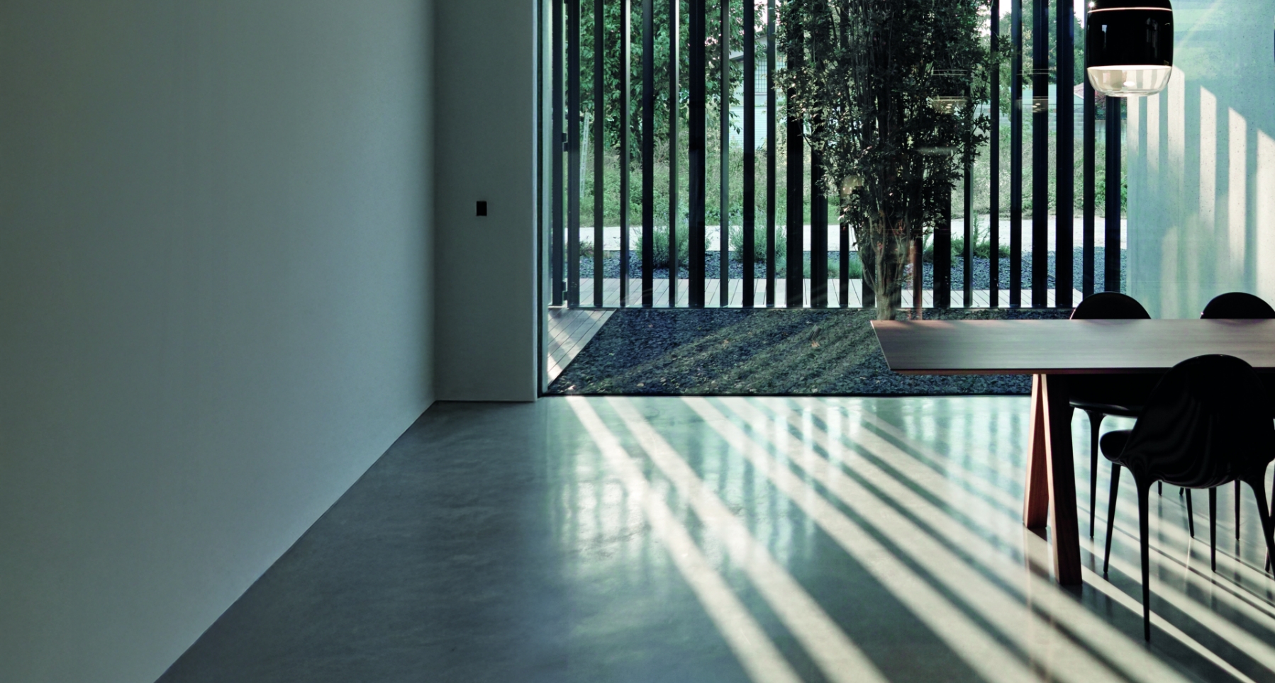 Luce naturale che attraversa grandi vetrate illumina pavimento in cemento sottile e continuo, tavolo moderno e sedie nere in ambiente minimalista.